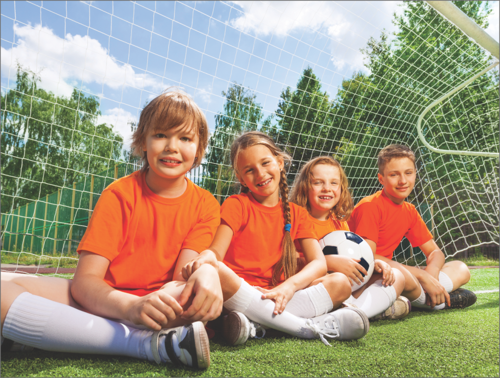 Children in orange soccer uniforms sitting on the grass, smiling and holding a soccer ball, with a goal net in the background, emphasizing teamwork and sportsmanship in children's activities.